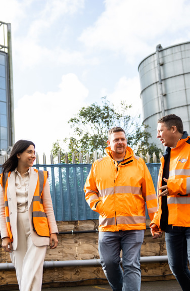 Three people in high vis jackets smiling on a sunny day and walking past the Energy from Waste plant. 