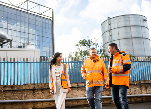 Three people in high vis jackets smiling on a sunny day and walking past the Energy from Waste plant.