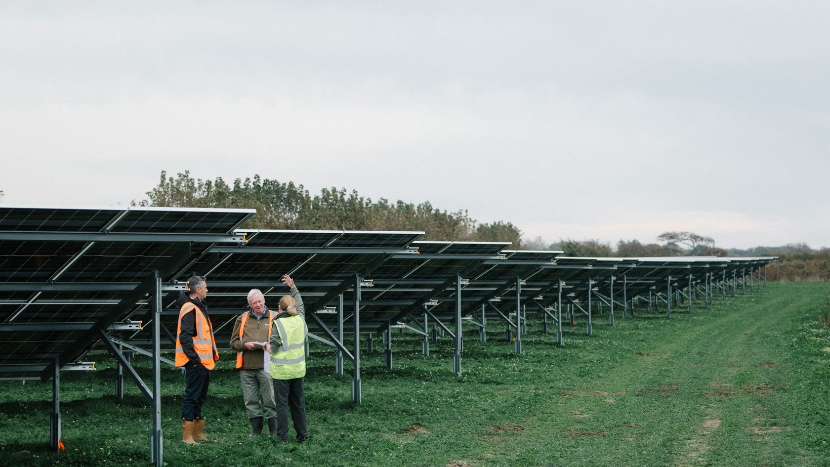Three people in high vis vests have a conversation standing in field underneath groung mount soalr panels