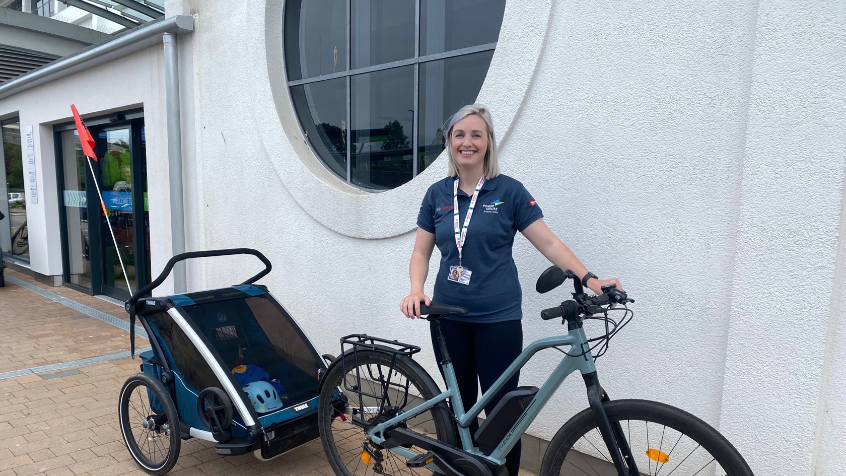 Sarah Naylor, Category Manager at the Powerhouse standing in a blue Powerhouse t-shirt next to her electric bike with Thule Chariot Cross trailer