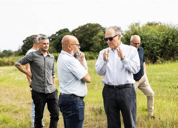 A photo of two men in white shirts and dark trousers deep in discussion. The man on the right is wearing sunglasses. The conversation is taking place in a green field. In the background are people talking and trees.