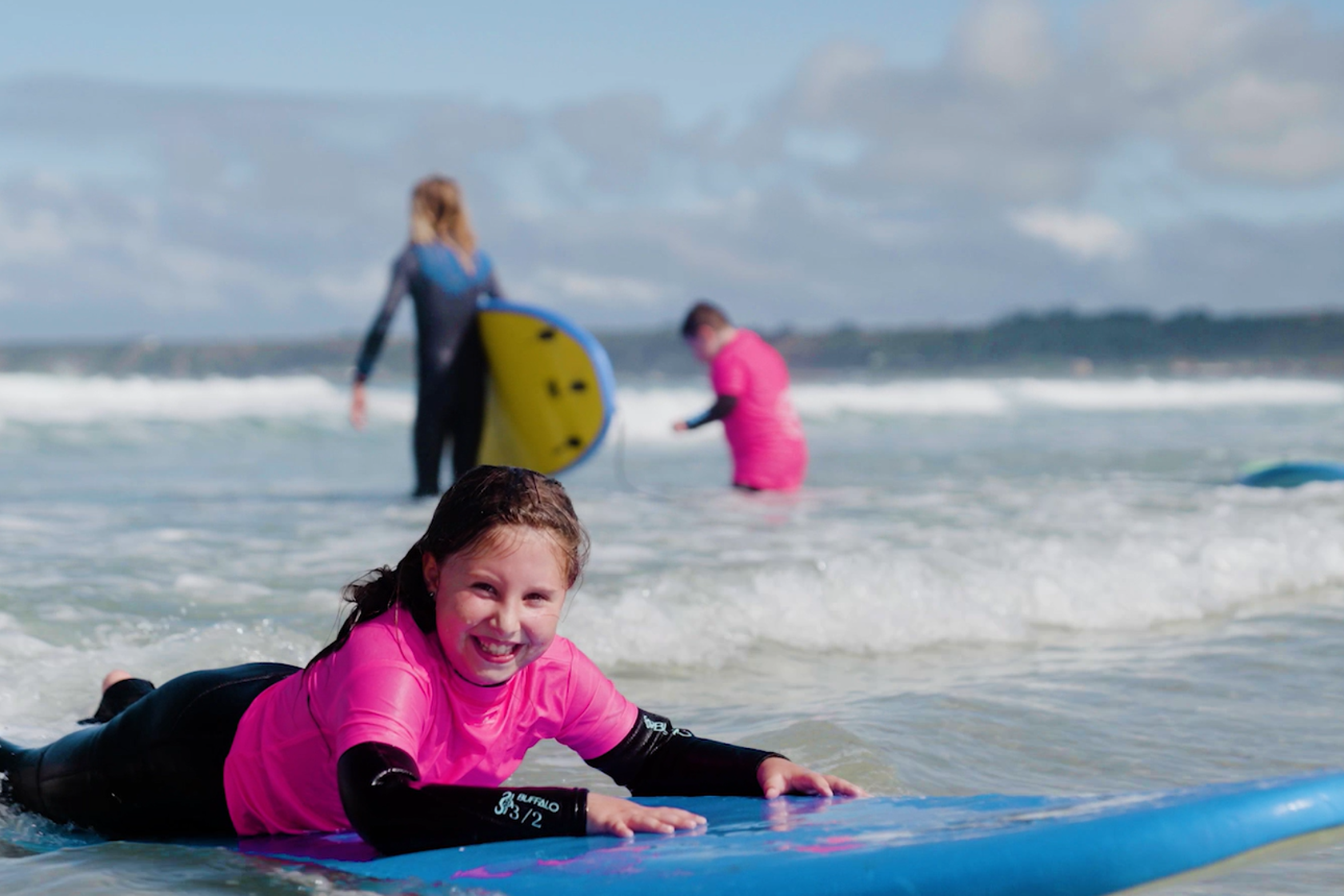 A child laying on a blue boogie board in the sea, smiling and wearing a pink top