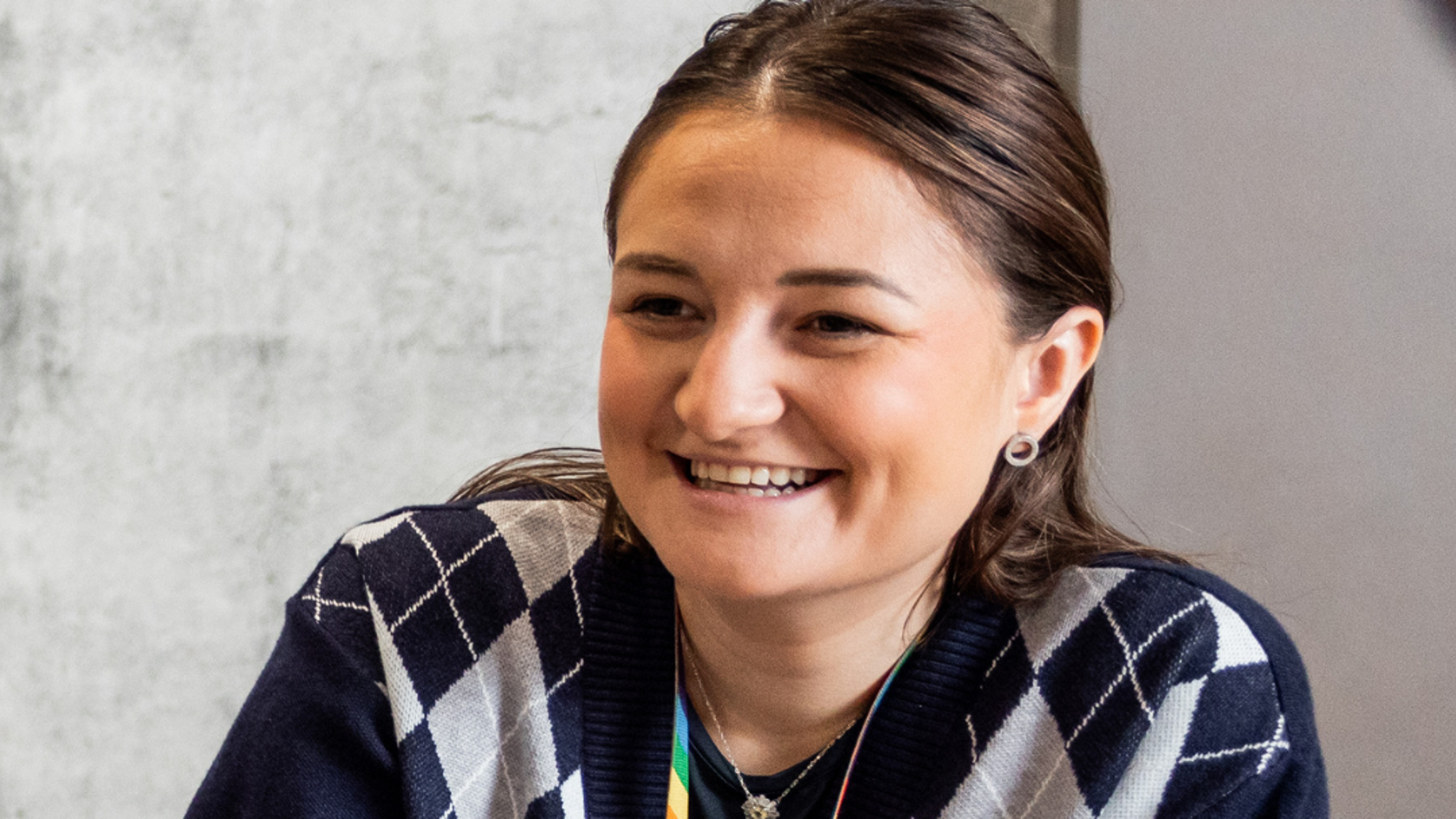 A person smiling, sat leaning on a meeting table wearing a blue cardigan