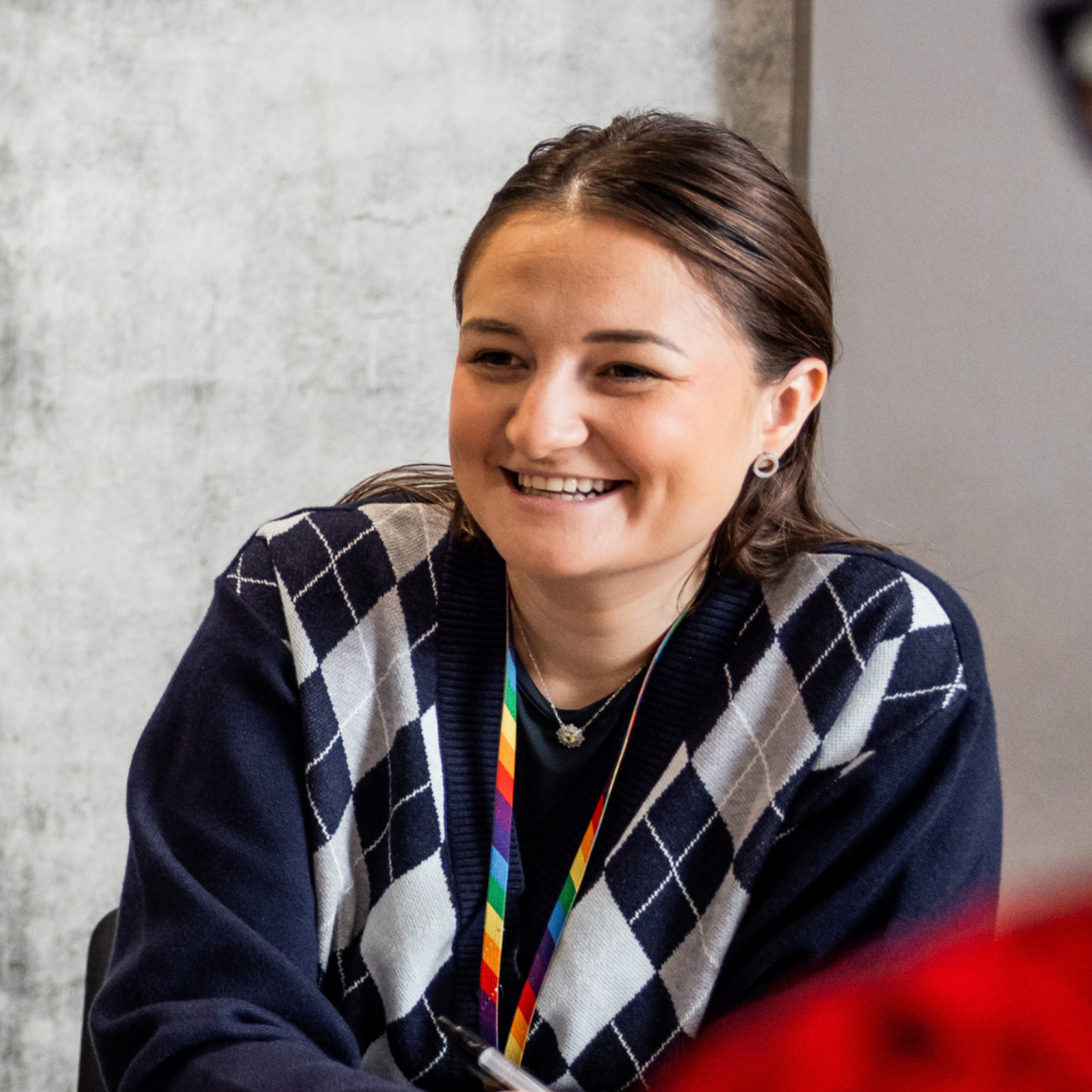 A person smiling, sat leaning on a meeting table wearing a blue cardigan