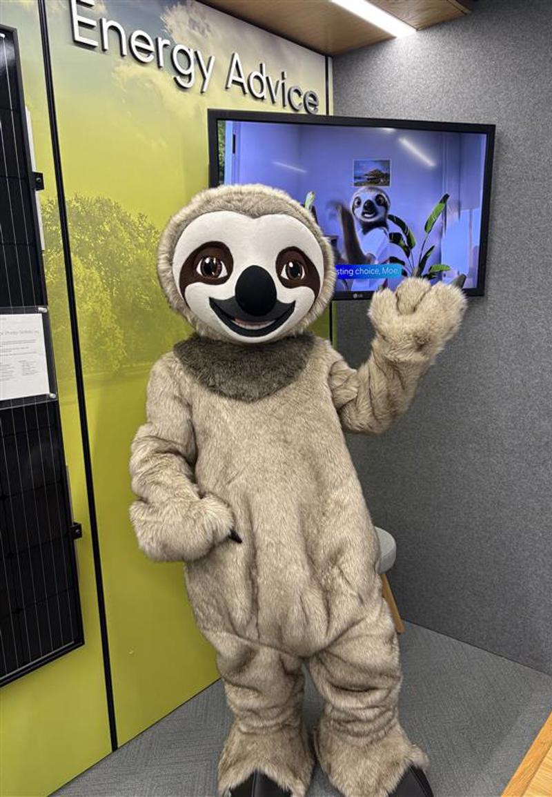 A person in a life size sloth suit, waving. Behind them is a TV screen featuring an animated sloth 