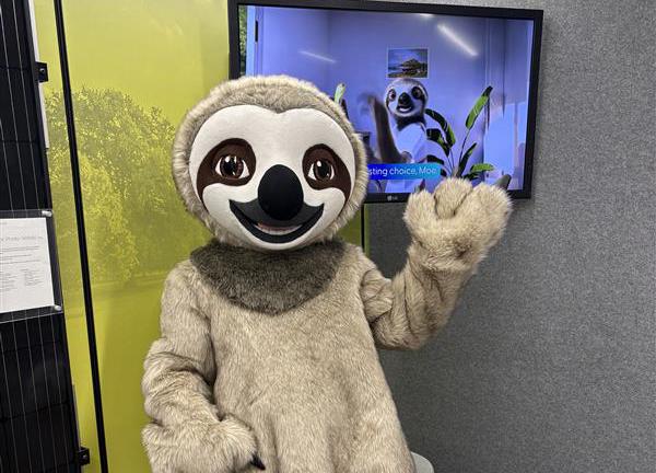 A person in a life size sloth suit, waving. Behind them is a TV screen featuring an animated sloth