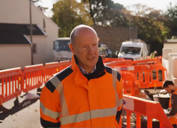 A person wearing orange high vis overalls on a sunny day, standing next next to roadworks
