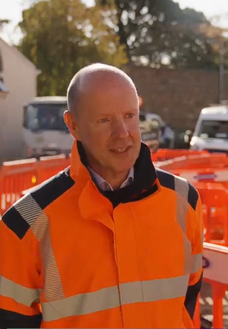 A person wearing orange high vis overalls on a sunny day, standing next next to roadworks