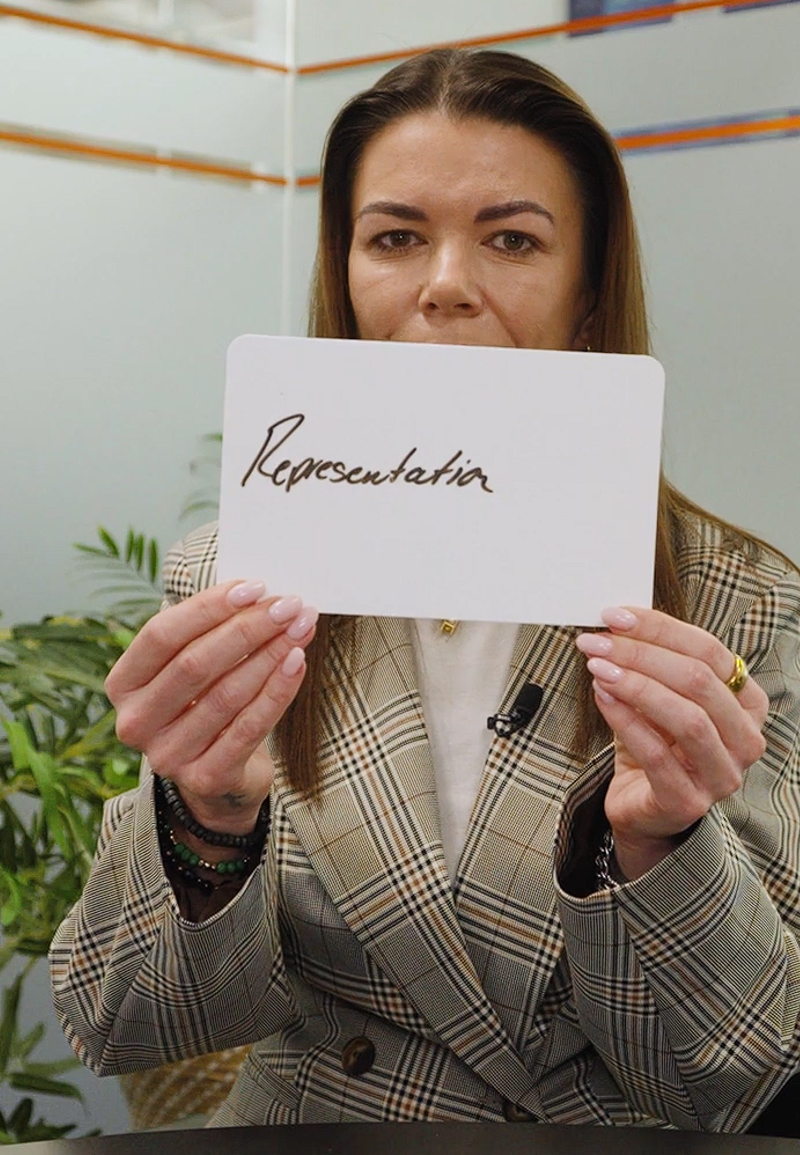 A person in an office setting holding a handwritten sign with the word 'representation'.