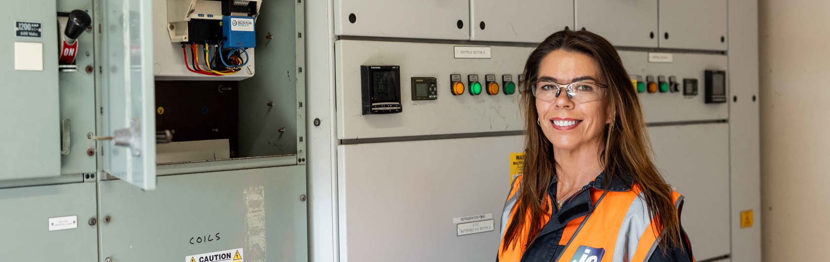 A woman in safety gear holds a tablet while standing in front of electrical control panels with warning labels.