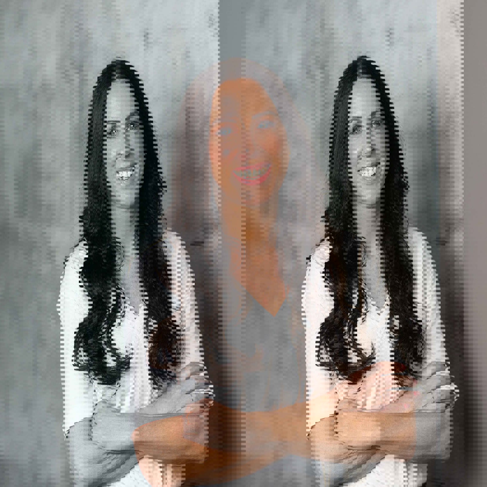 A lady wearing a white t-shirt with long wavy, brown hear standing against a grey background with her arms folded.