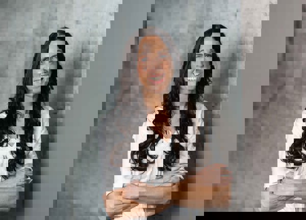 A lady wearing a white t-shirt with long wavy, brown hear standing against a grey background with her arms folded.