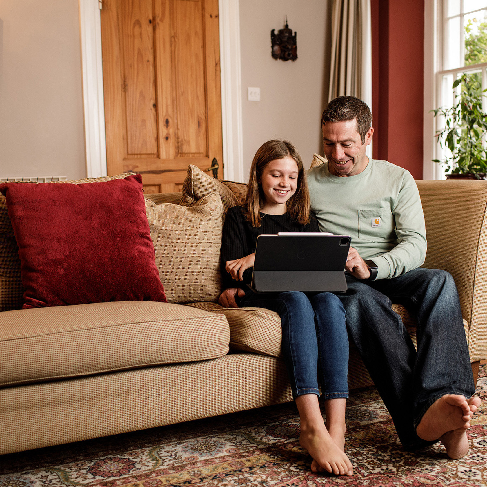 Father and daughter sit on a sofa whilst enjoying using a tablet.