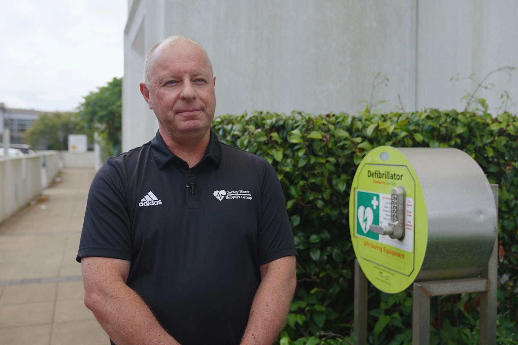 A person in a blue t-shirt standing outside a building, next to a defibullator