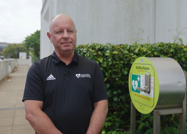 A person in a blue t-shirt standing outside a building, next to a defibullator