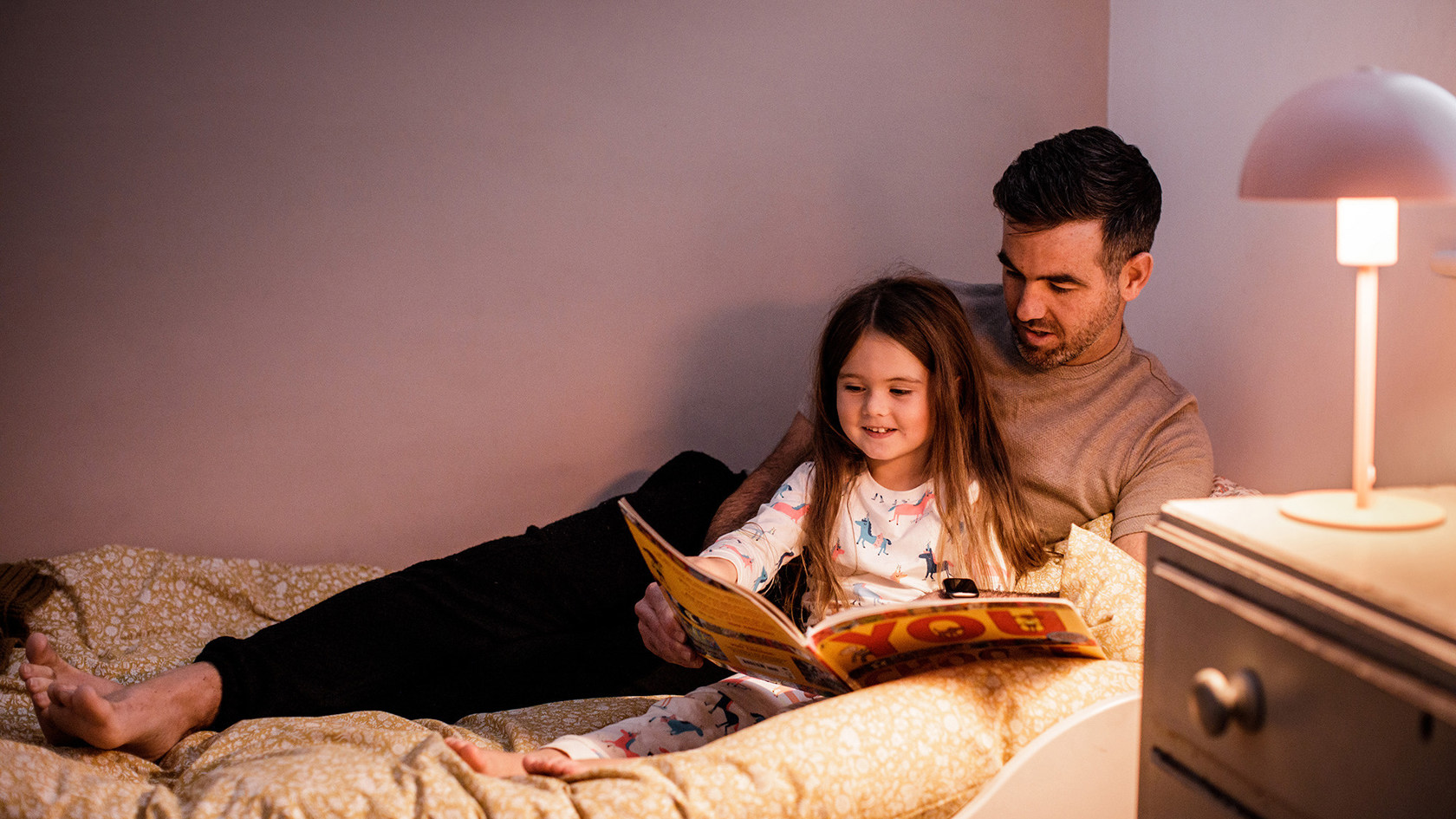 A father reads a story to his daughter in bed.